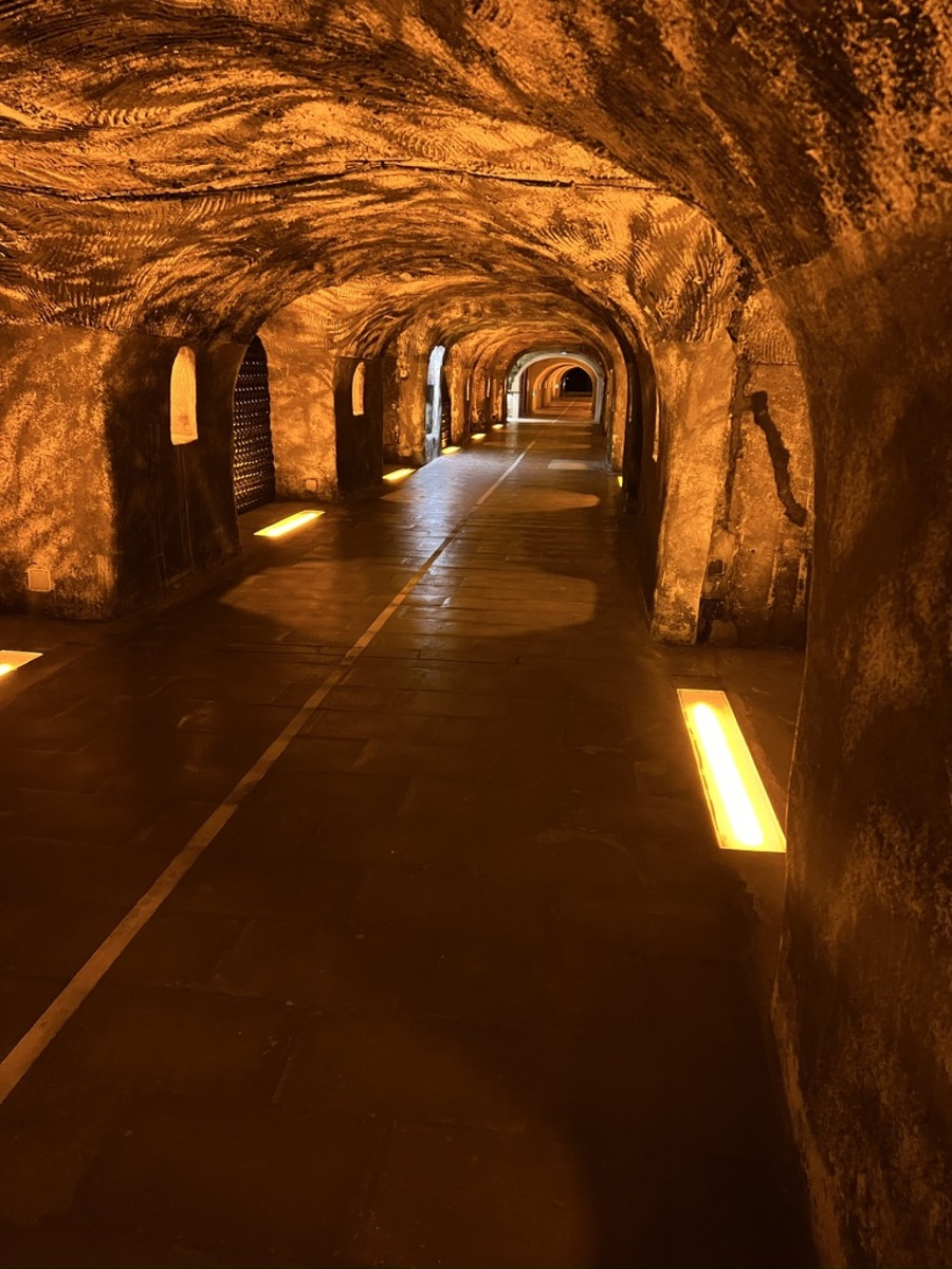 Dimly lit tunnel with textured, arching walls and a tiled floor, illuminated in warm, amber tones. A faint line runs down the center, creating a mysterious, inviting atmosphere.