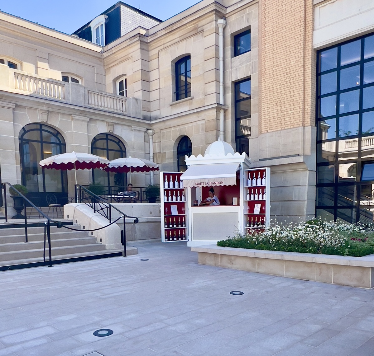 Elegant courtyard with a Moët & Chandon kiosk, surrounded by a grand building. Red-and-white umbrellas provide shade to seating on the steps.