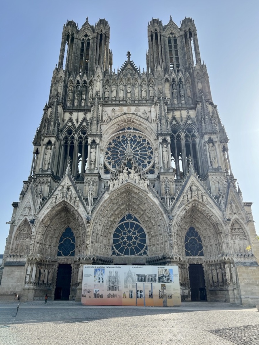 Reims Notre Dame cathedral facade with intricate stone carvings, three large doorways, and a prominent rose window. Sunlit exterior conveys historic grandeur.