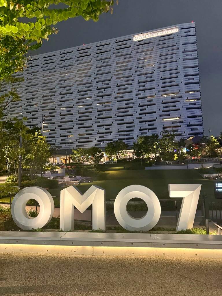 Illuminated "OMO7" sign in a park-like setting at night, with a modern, patterned building in the background. The scene feels serene and urban.