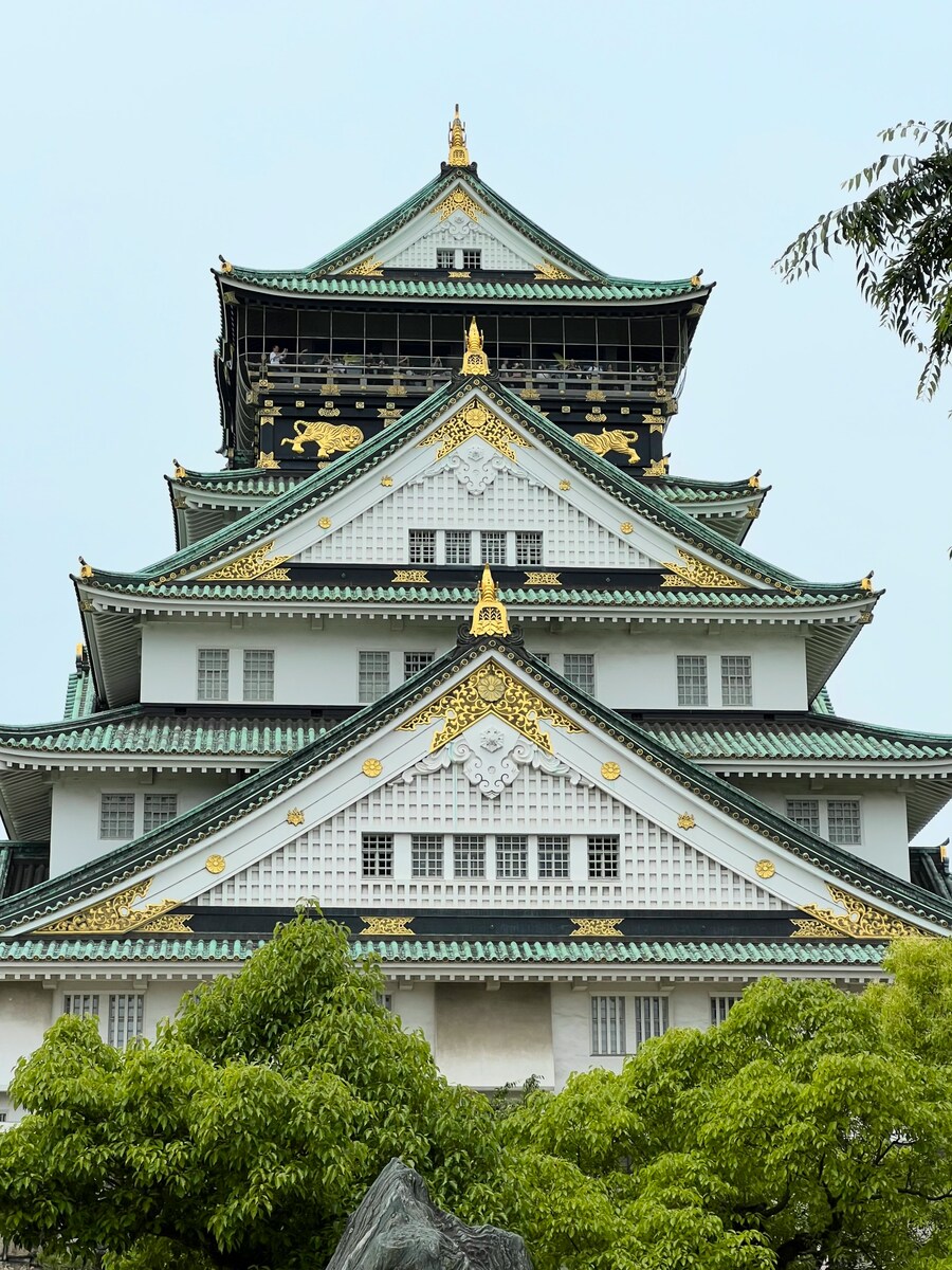 A majestic Japanese castle with green roofs and gold accents stands under a clear sky, framed by lush green trees. The scene conveys grandeur and serenity.