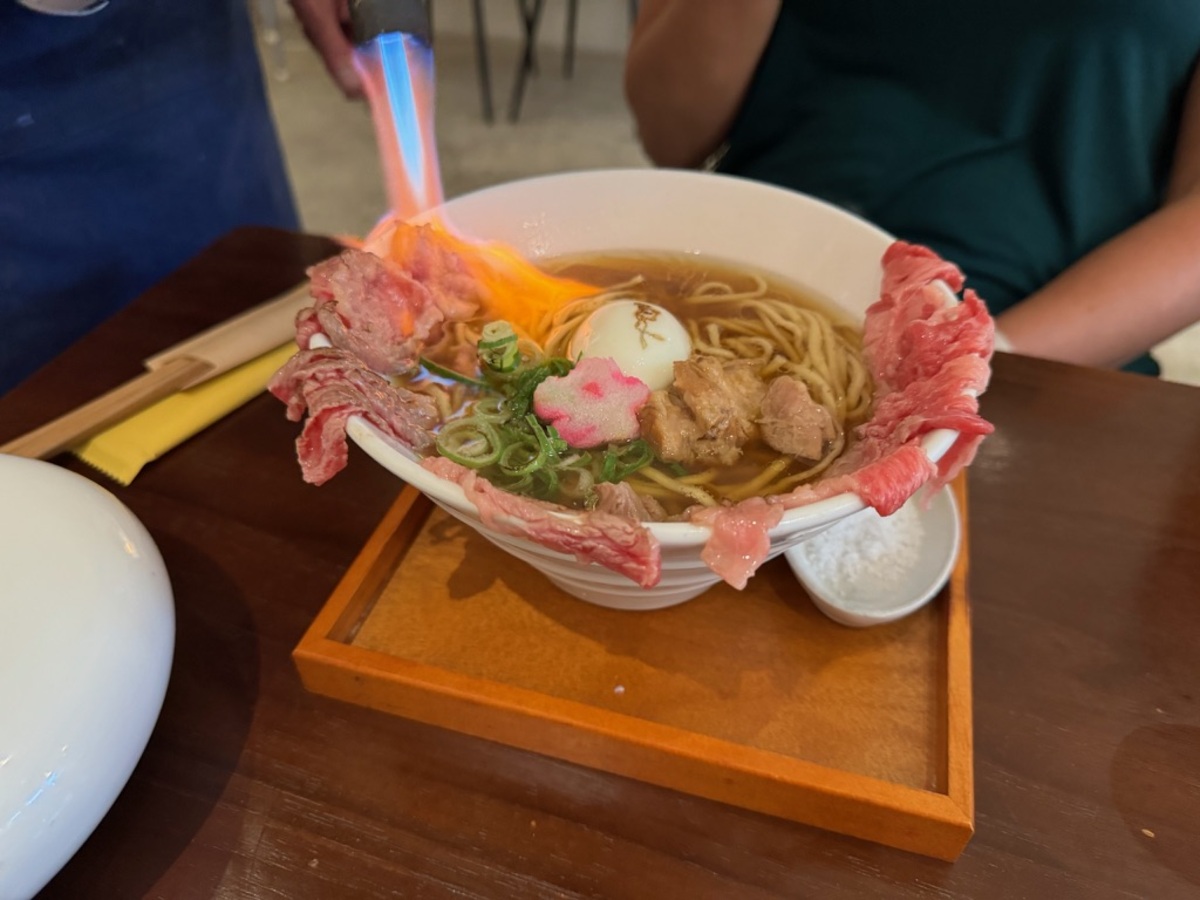 A bowl of ramen topped with raw beef slices, being torched to cook. The dish includes a soft-boiled egg, green onions, and a decorative pink element.