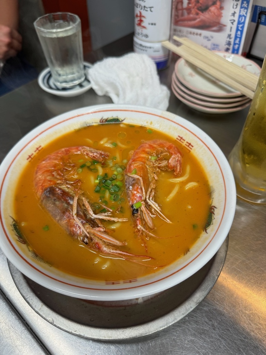 A bowl of spicy noodle ramen with two whole shrimp, garnished with chopped green onions, is on a table. Nearby are chopsticks and a glass of water.