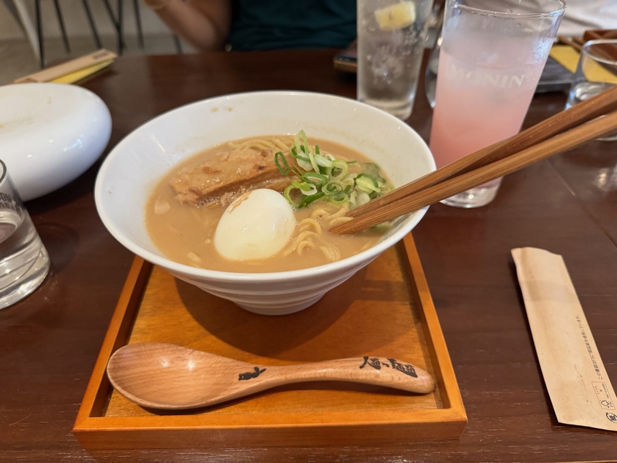 A bowl of ramen with sliced pork, a soft-boiled egg, and green onions, served on a wooden tray. A pink drink and chopsticks are beside it. Cozy, inviting setting.