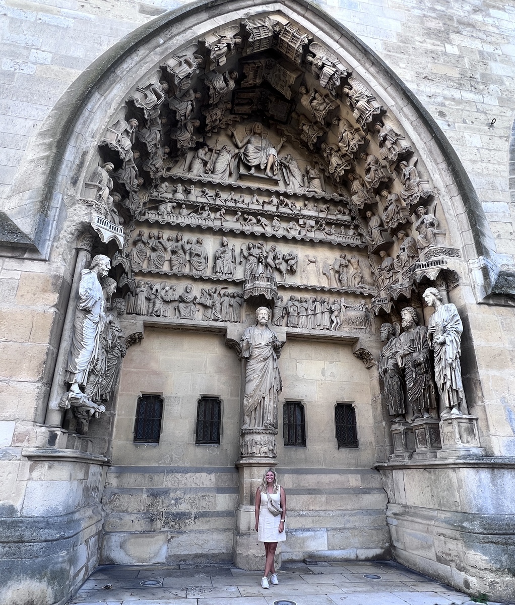 Stone cathedral facade featuring intricate, detailed religious sculptures depicting biblical scenes. A person in a light dress stands at the base, highlighting the structure's impressive scale. The tone is awe-inspiring and historic.