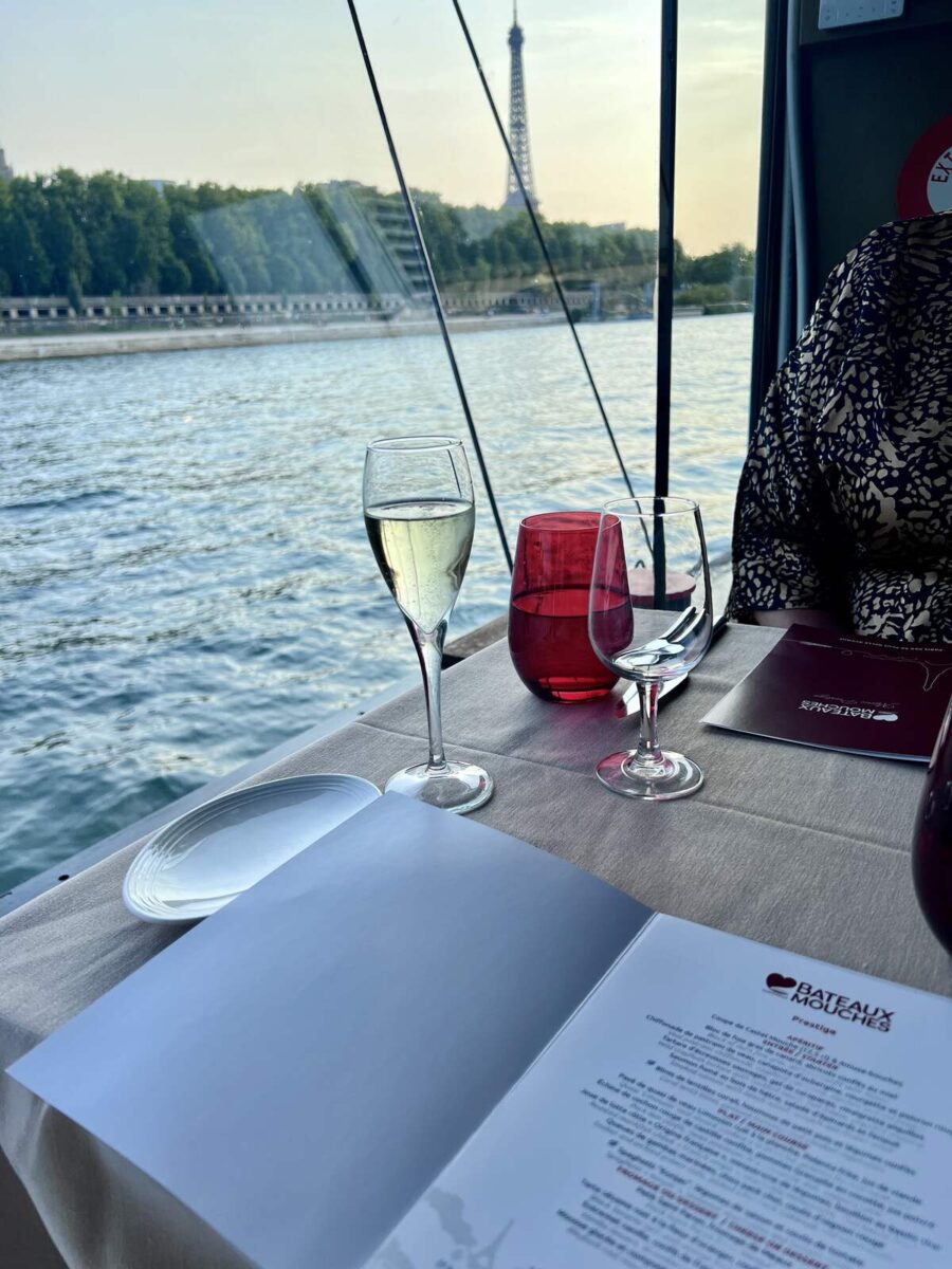 A riverside dining table with a menu, wine glasses, and red candle holders. The Eiffel Tower and calm water are in the background, evoking a serene, elegant ambiance.