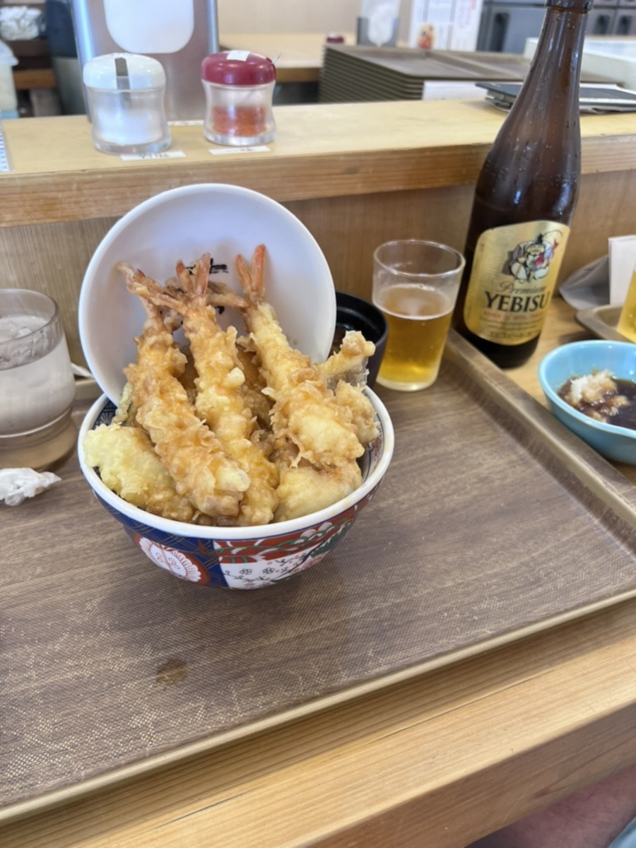 A bowl of golden, crispy tempura shrimp with assorted vegetables sits on a tray. A bottle and glass of beer is beside it, creating a casual dining atmosphere.