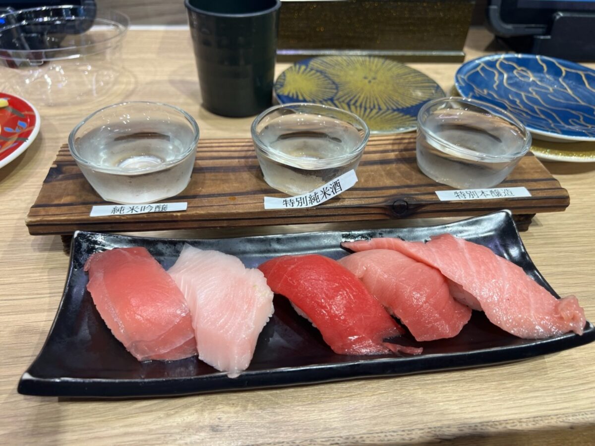 A black plate with five pieces of sushi, featuring pink and red fish, sits in front of three glass sake cups on a wooden board. Elegant and inviting.
