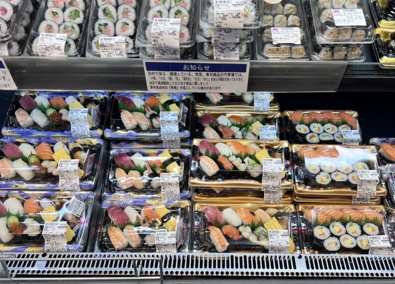 A supermarket shelf filled with a variety of sushi trays. Vibrant rolls and nigiri of different colors and ingredients are neatly arranged in plastic packaging.