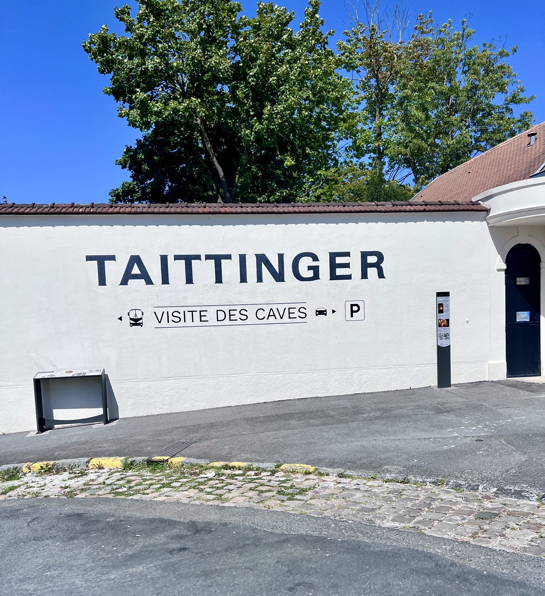 White wall with "TAITTINGER" in bold black letters, signage indicating "VISITE DES CAVES" with arrows, trees in the background, blue sky above.