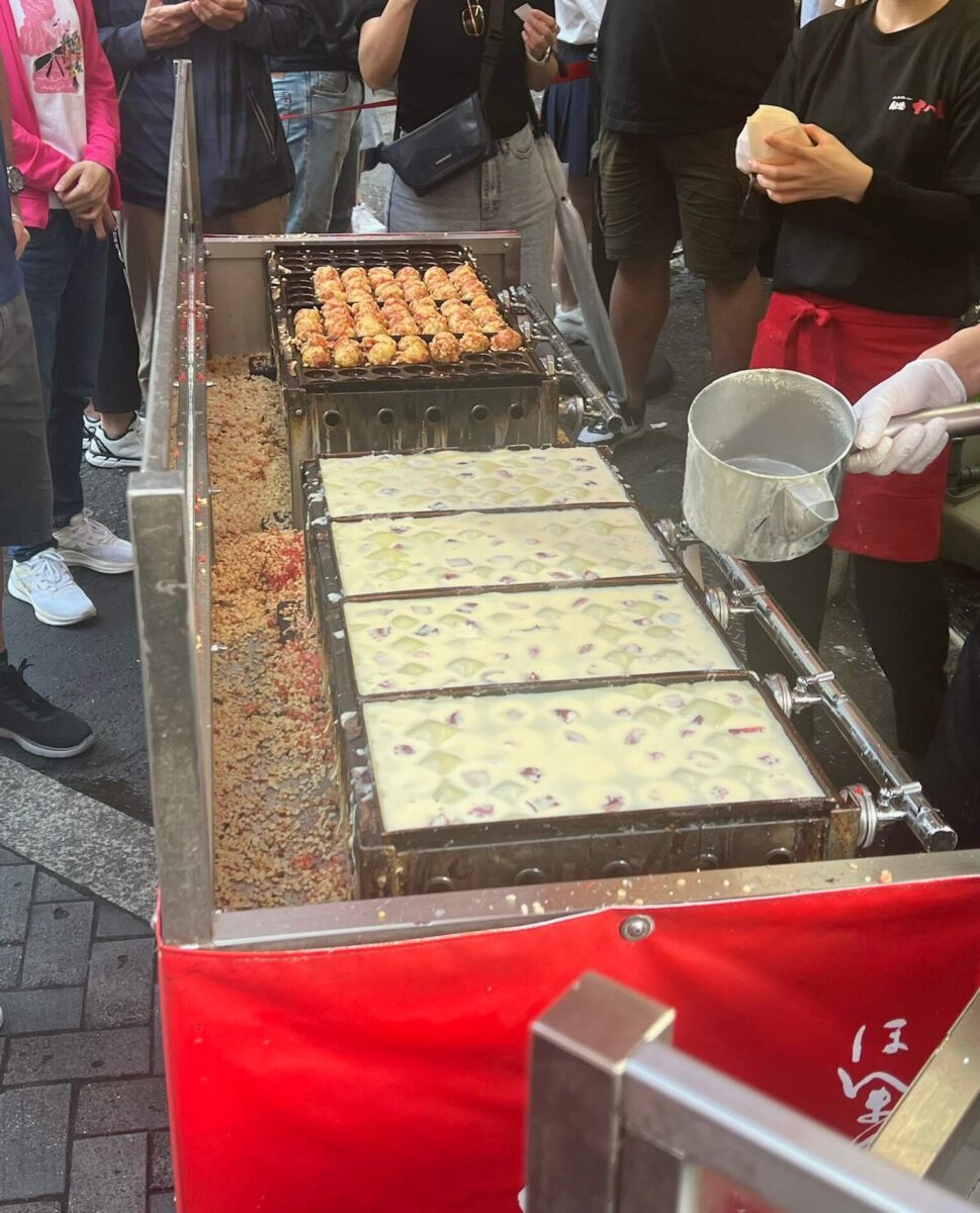 Street vendor making takoyaki on a hot grill. Raw batter fills molds beside cooked golden balls. Crowd eagerly watches the cooking process. Energetic scene.