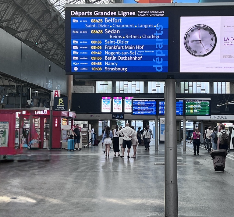 Train station interior with a large departures board displaying destinations and times, including Belfort and Sedan. Travelers walk below, creating a bustling atmosphere.