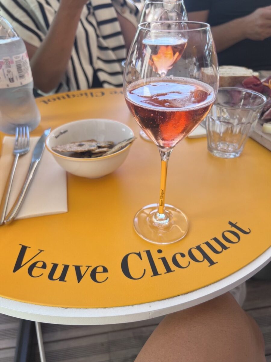 A glass of rosé champagne sits on a vibrant yellow table with "Veuve Clicquot" text. Nearby, there are a bowl, utensils, and water, suggesting a relaxed dining setting.