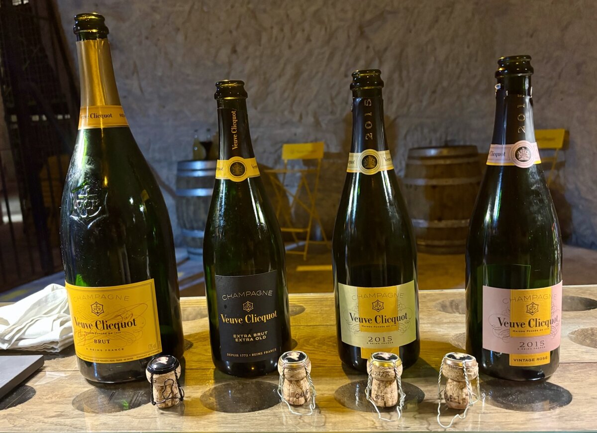 Four Veuve Clicquot champagne bottles of different styles and sizes on a wooden table, with corks and metal wire cages in front, in a rustic setting.