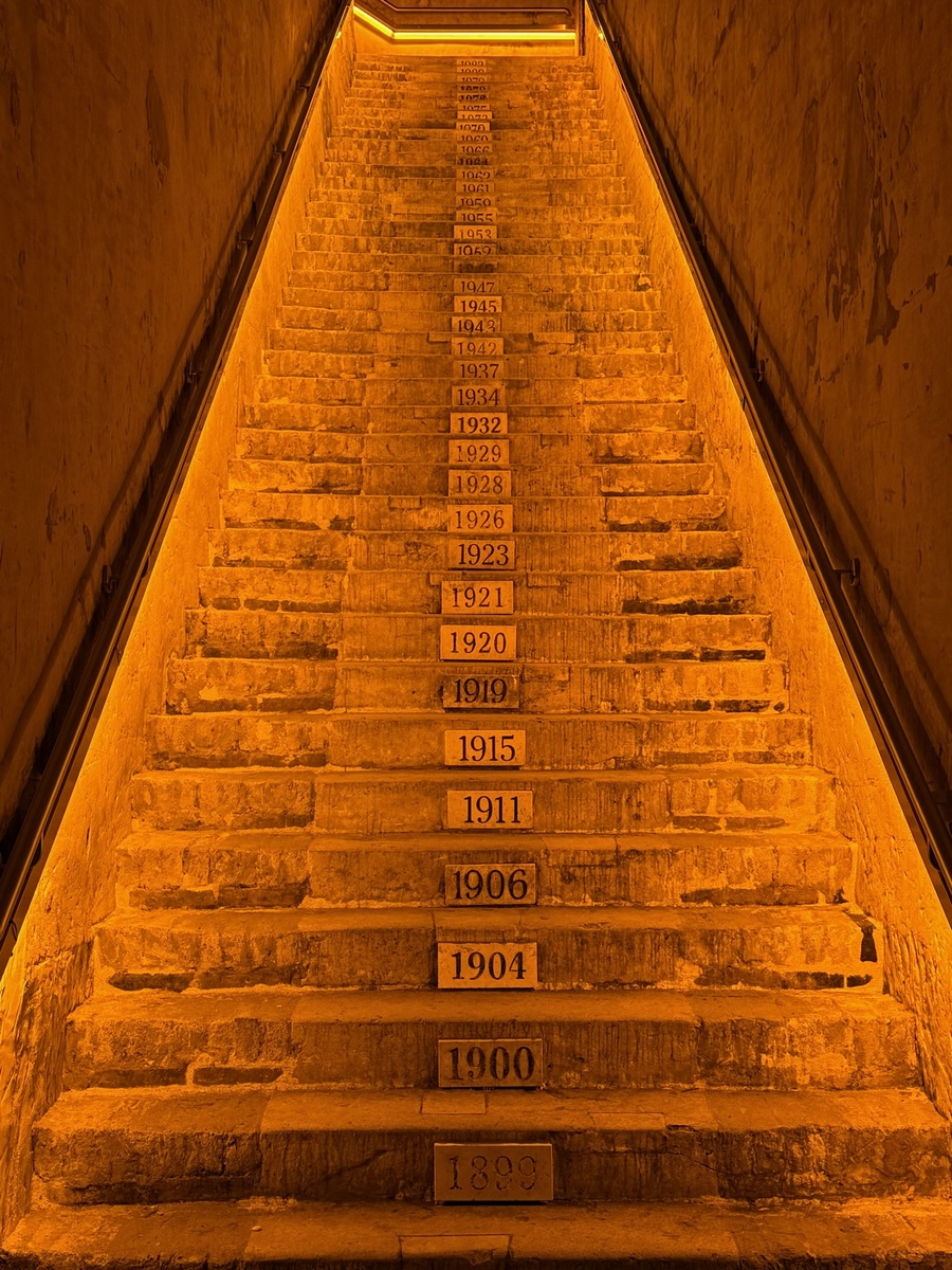 Illuminated staircase at Veuve Clicquot in Reims with years 1889 to 1947 marked on steps, creating a historical timeline. Warm orange lighting adds a dramatic, nostalgic feel.