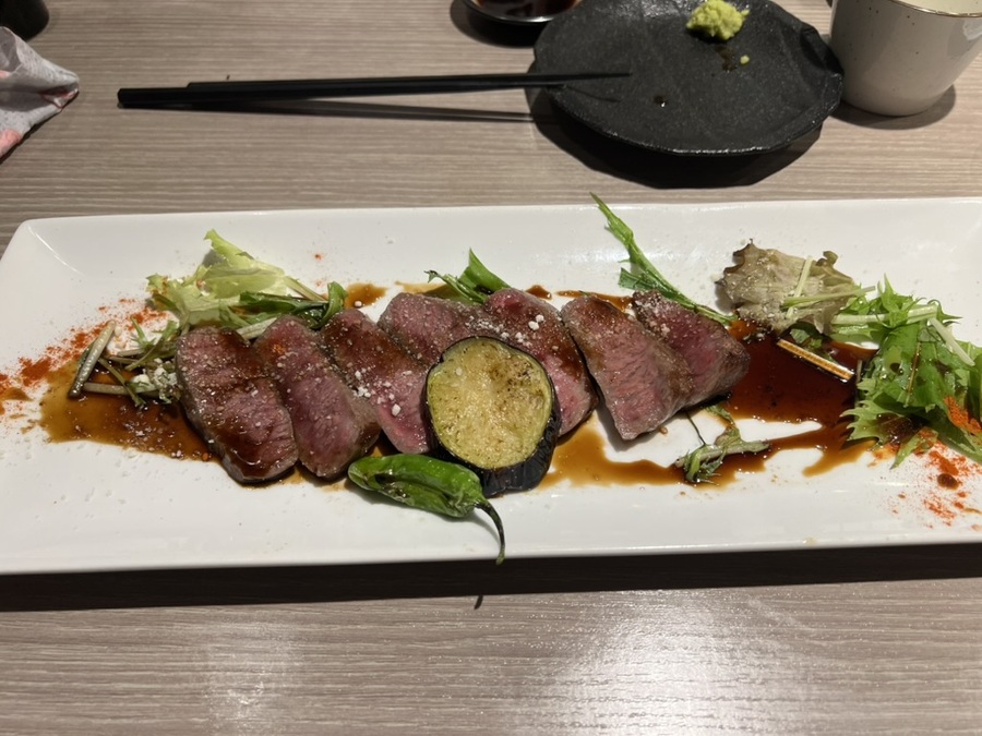Sliced rare A5 steak with seared eggplant on a rectangular plate, garnished with mixed greens and sauce. A small black dish and chopsticks nearby.