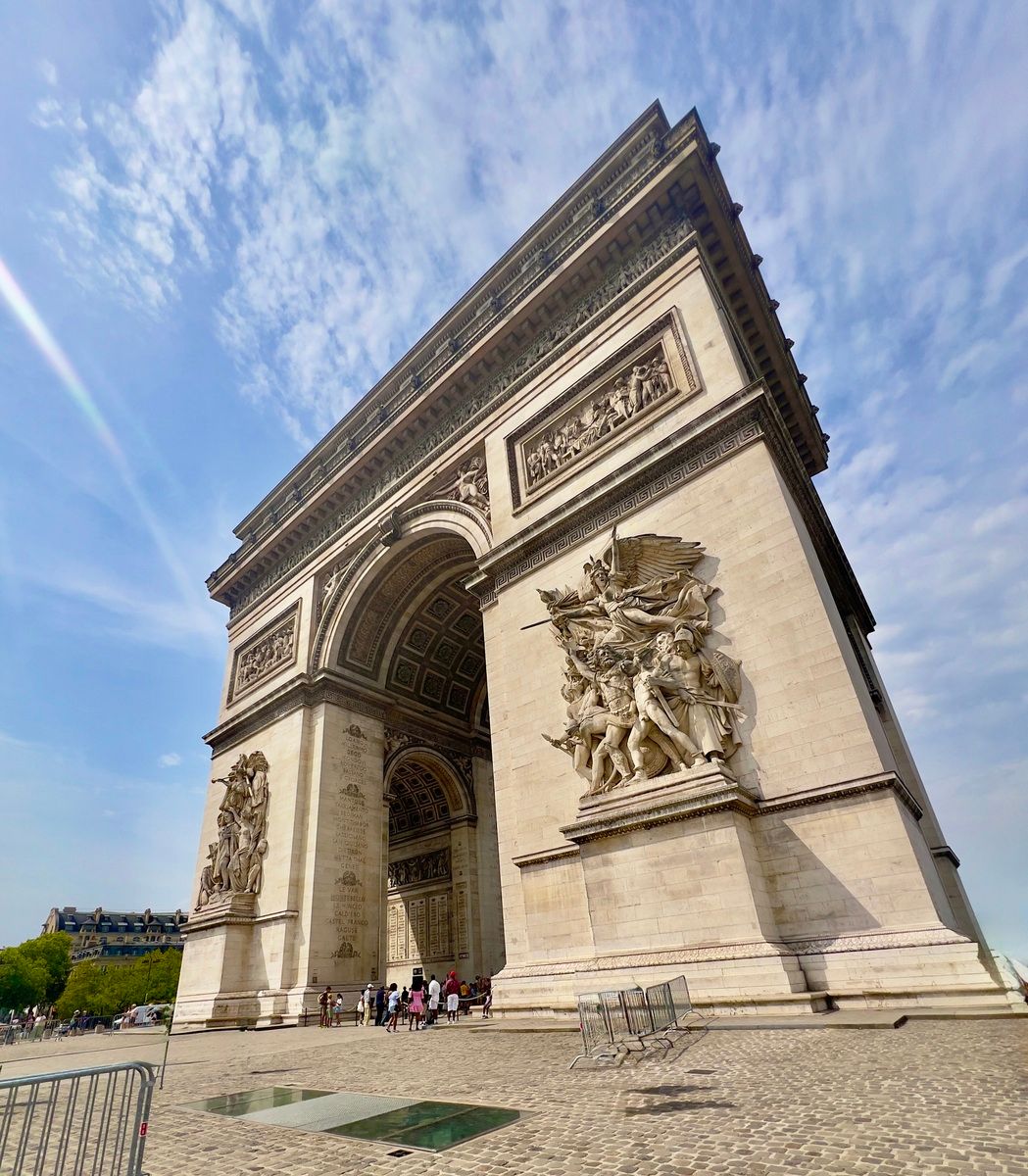 The image shows the Arc de Triomphe in Paris, viewed from below against a partly cloudy sky. Tourists gather at its base, and detailed sculptures adorn the arch.