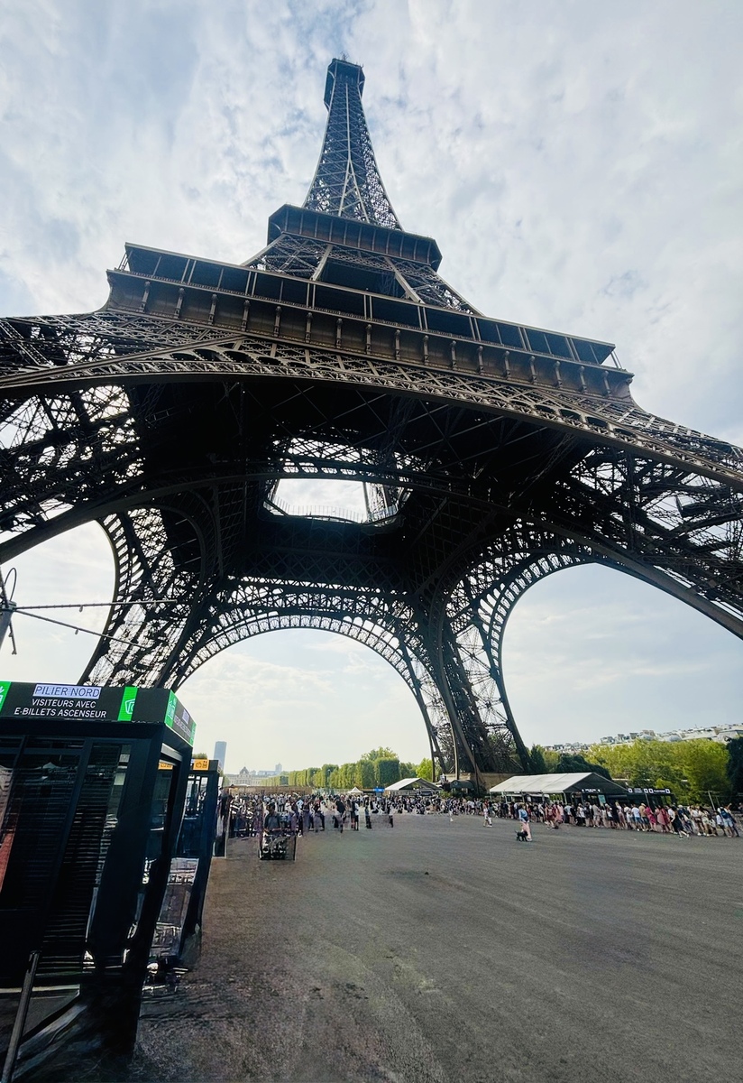 View from beneath the Eiffel Tower against a partly cloudy sky. Tourists gather below; ticket booth visible to the left. Energetic and iconic atmosphere.