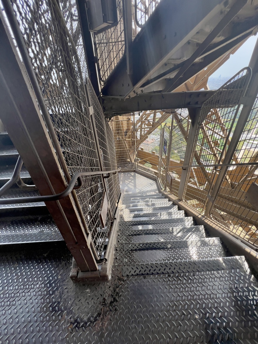 Metal staircase with mesh railings and sunlight streaming through. The steps lead downwards, surrounded by metal beams, suggesting an industrial setting.