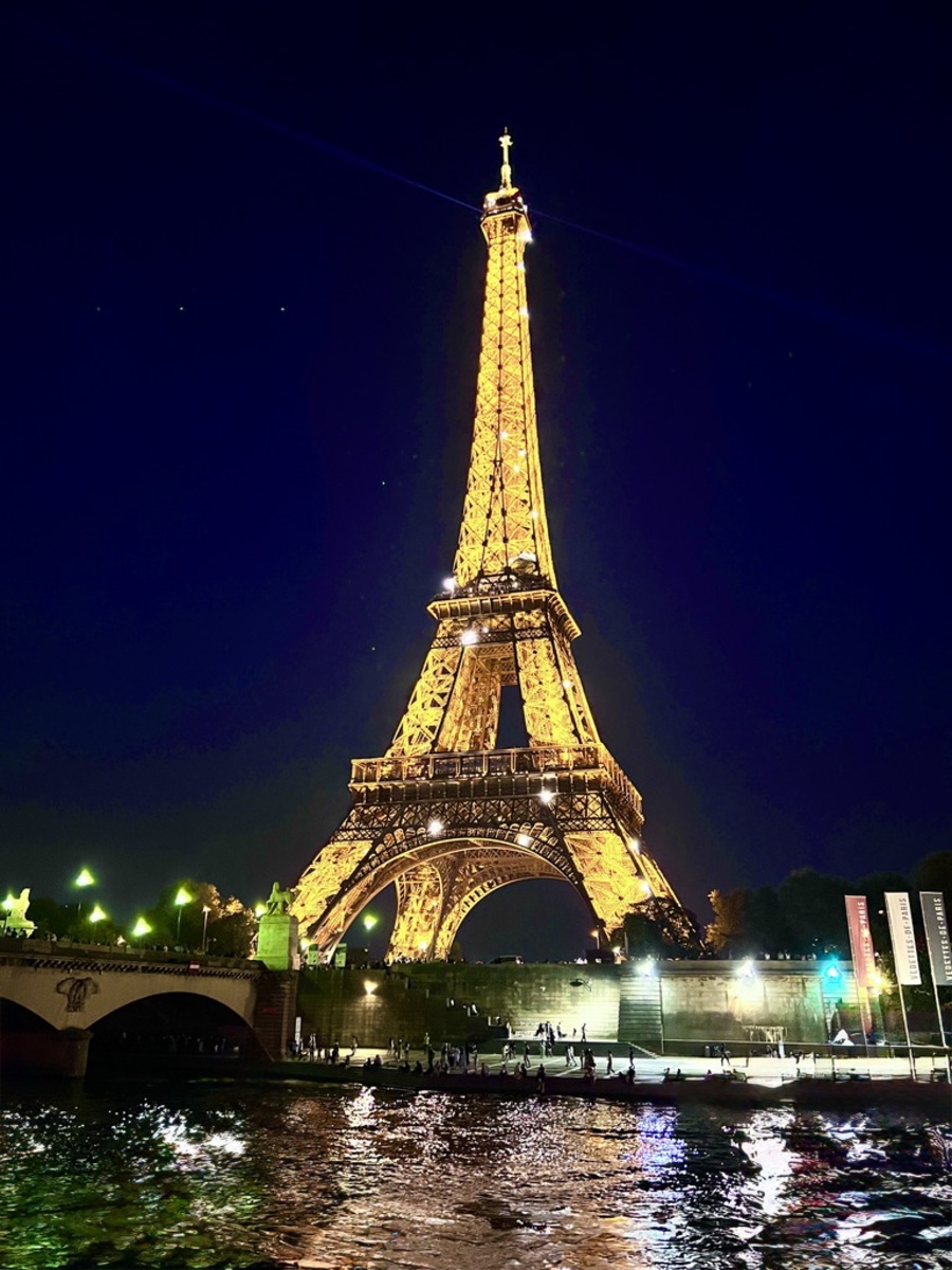 Illuminated Eiffel Tower at night, glowing gold against a dark blue sky. Reflections shimmer in the Seine River, creating a romantic, enchanting scene.