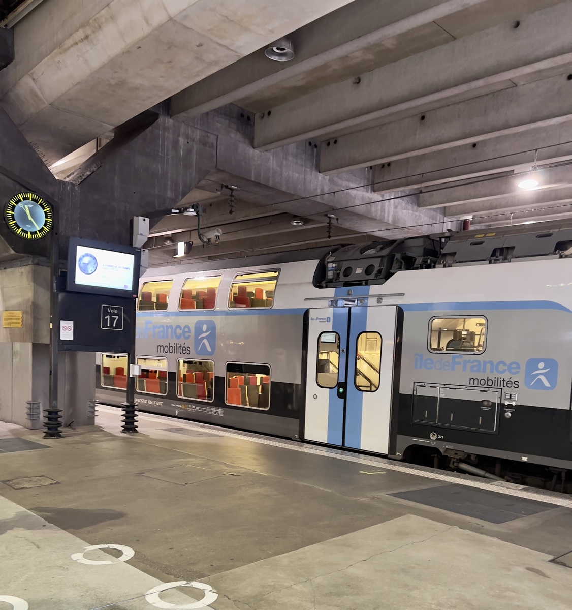 Double-decker train at an urban station with concrete beams overhead. A clock and digital sign are visible, displaying platform 17. Quiet and modern atmosphere.