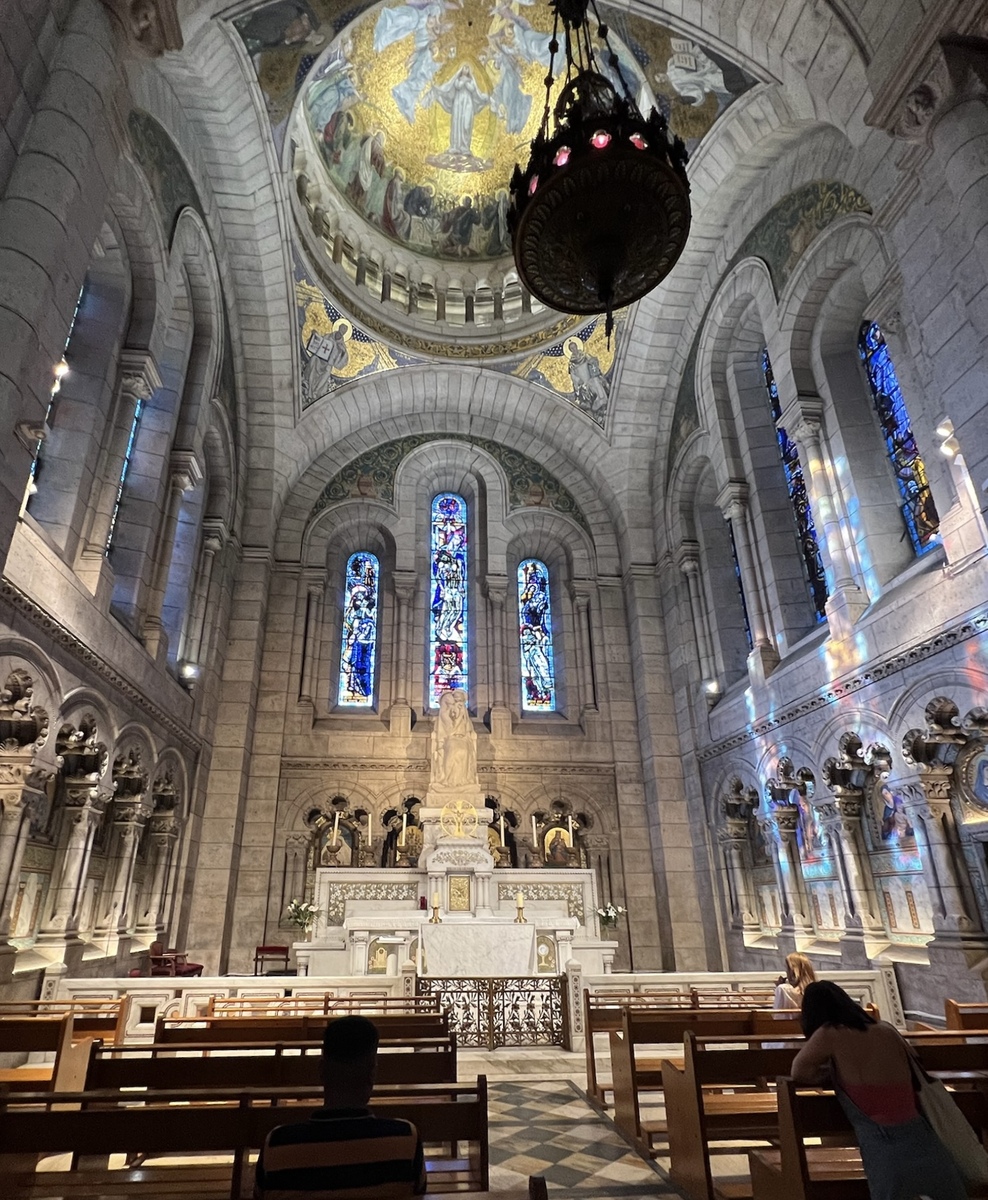 Interior of a grand cathedral with a high arched ceiling and a large chandelier. Stained-glass windows and a detailed altar create a reverent atmosphere.