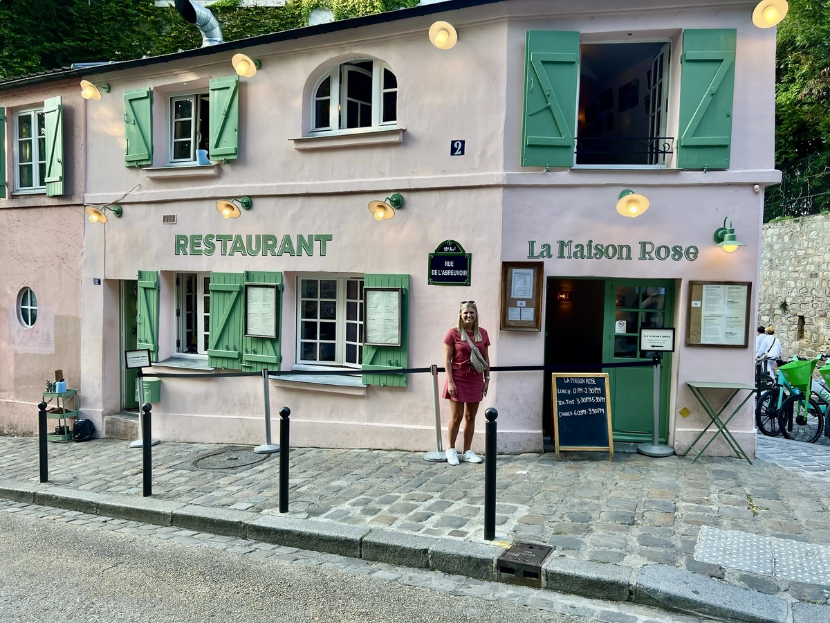 Brenda stands outside La Maison Rose, a quaint pink restaurant with green shutters in Montmartre, Paris. The setting is charming and inviting.