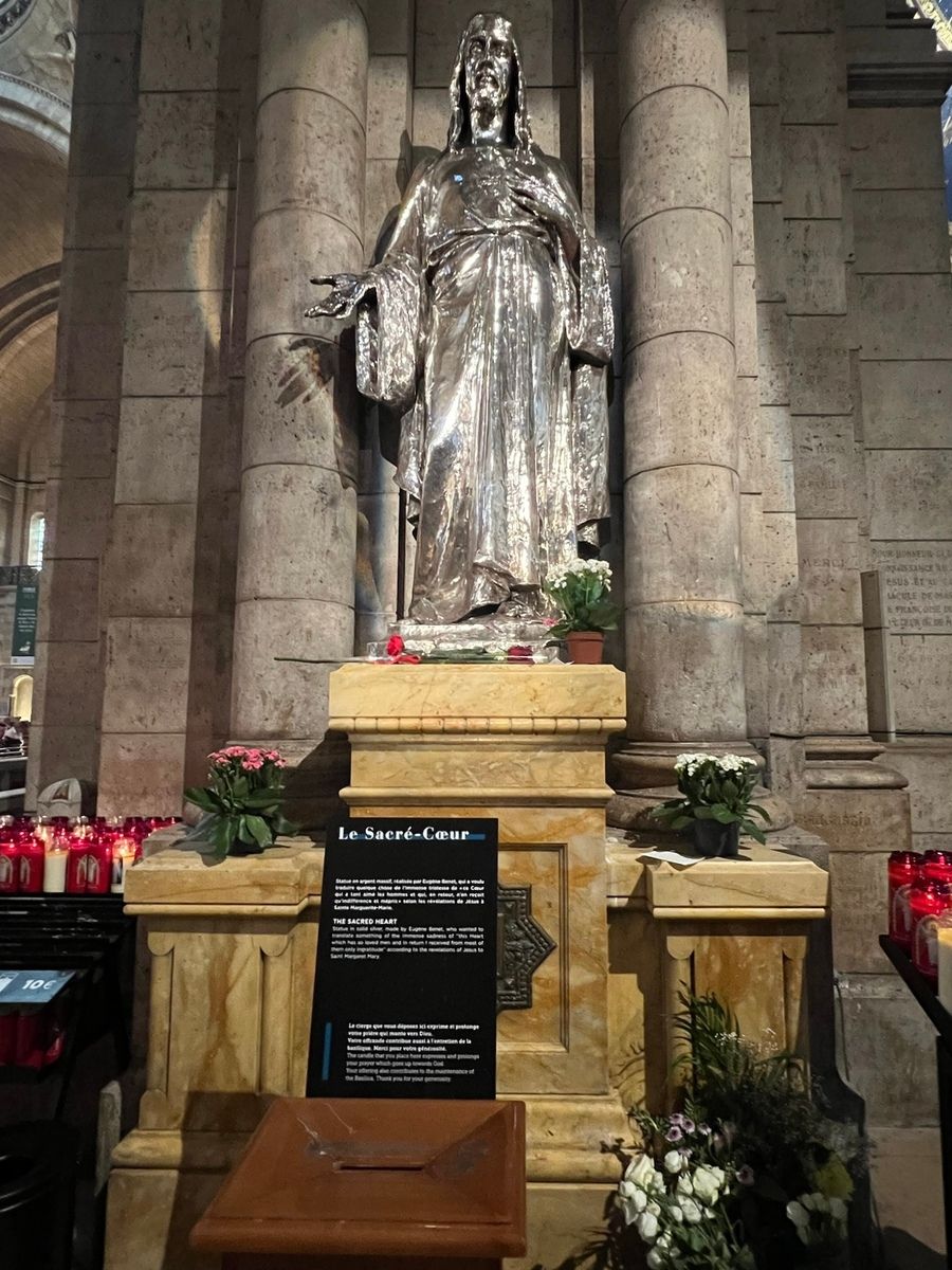 Statue of standing Jesus with open arms, surrounded by lit candles and flowers, set against a stone wall. The setting feels solemn and venerable.
