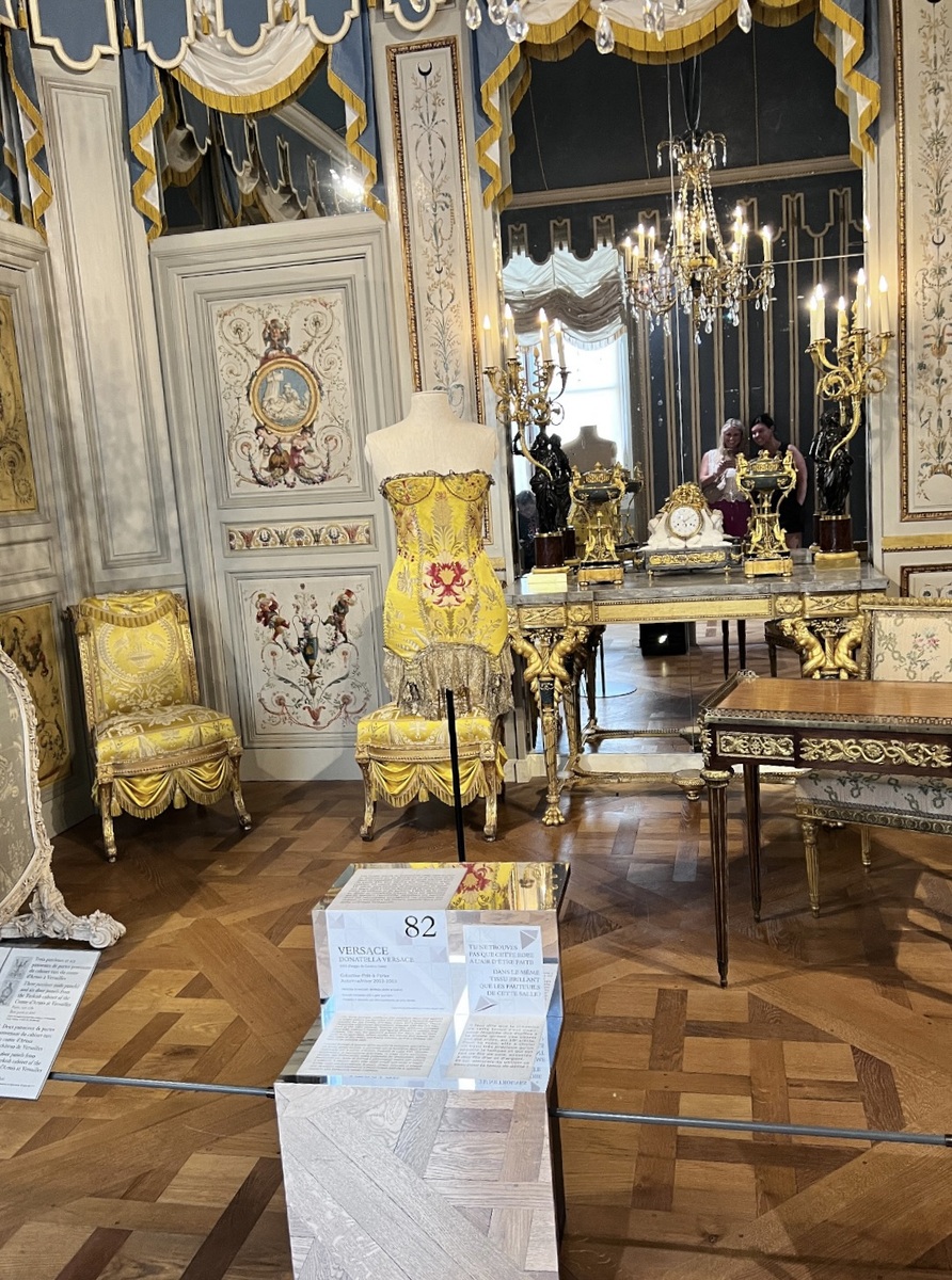 Luxurious room with ornate decor, featuring a Versace yellow embroidered dress on a mannequin. Gilded furnishings, vintage clock, and chandelier add elegance.