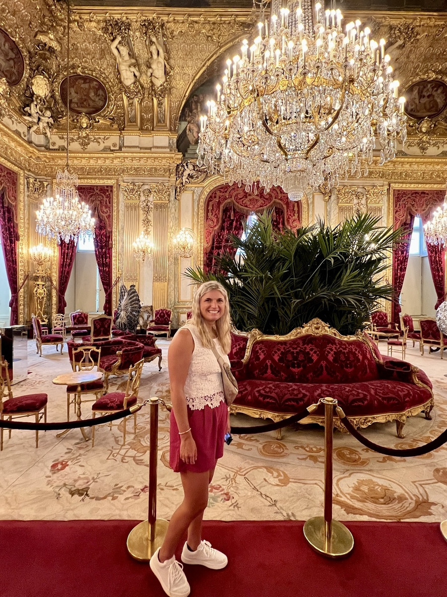 A woman smiles in an opulent room featuring ornate gold chandeliers, red velvet furniture, and lavish decor. The atmosphere is luxurious and grand.