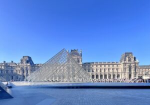 The Louvre pyramid stands prominently in front of a historic stone building, under a bright blue sky, surrounded by people and exuding a sense of grandeur and modernity.