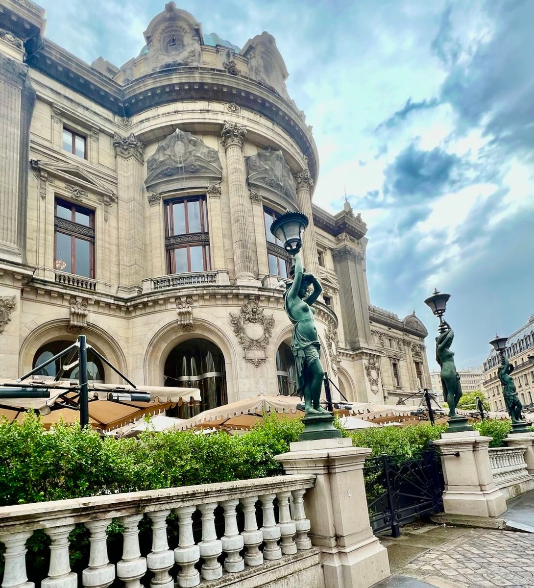 Opera Garnier in Paris with tall arched windows, detailed stone carvings, and decorative statues holding lamps. Overcast sky, creating a dramatic tone.