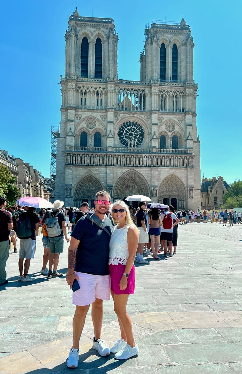 A smiling couple stands in front of Notre-Dame Cathedral on a sunny day. The historic facade is detailed and the plaza is crowded with tourists.