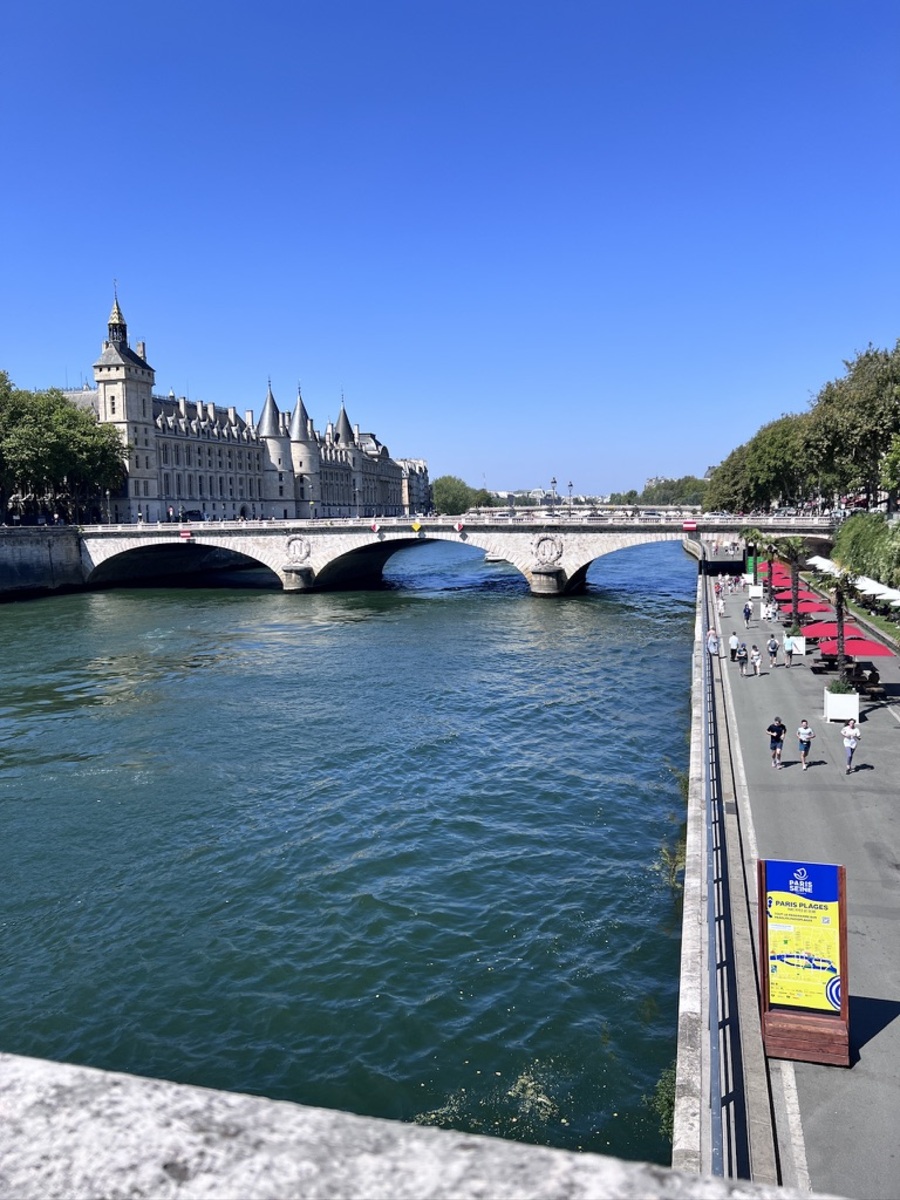 A scenic view of the Seine River with a historic stone bridge under a clear blue sky. People stroll along the riverside walkway lined with trees and red canopies, evoking a tranquil and leisurely atmosphere in a bustling city.