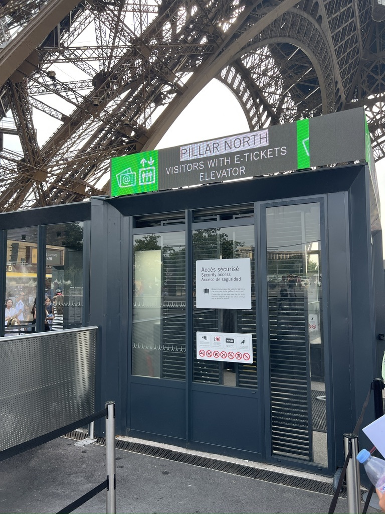 Entrance to the Eiffel Tower’s Pillar North elevator for e-ticket holders. The modern structure contrasts with the iconic tower's intricate ironwork above.