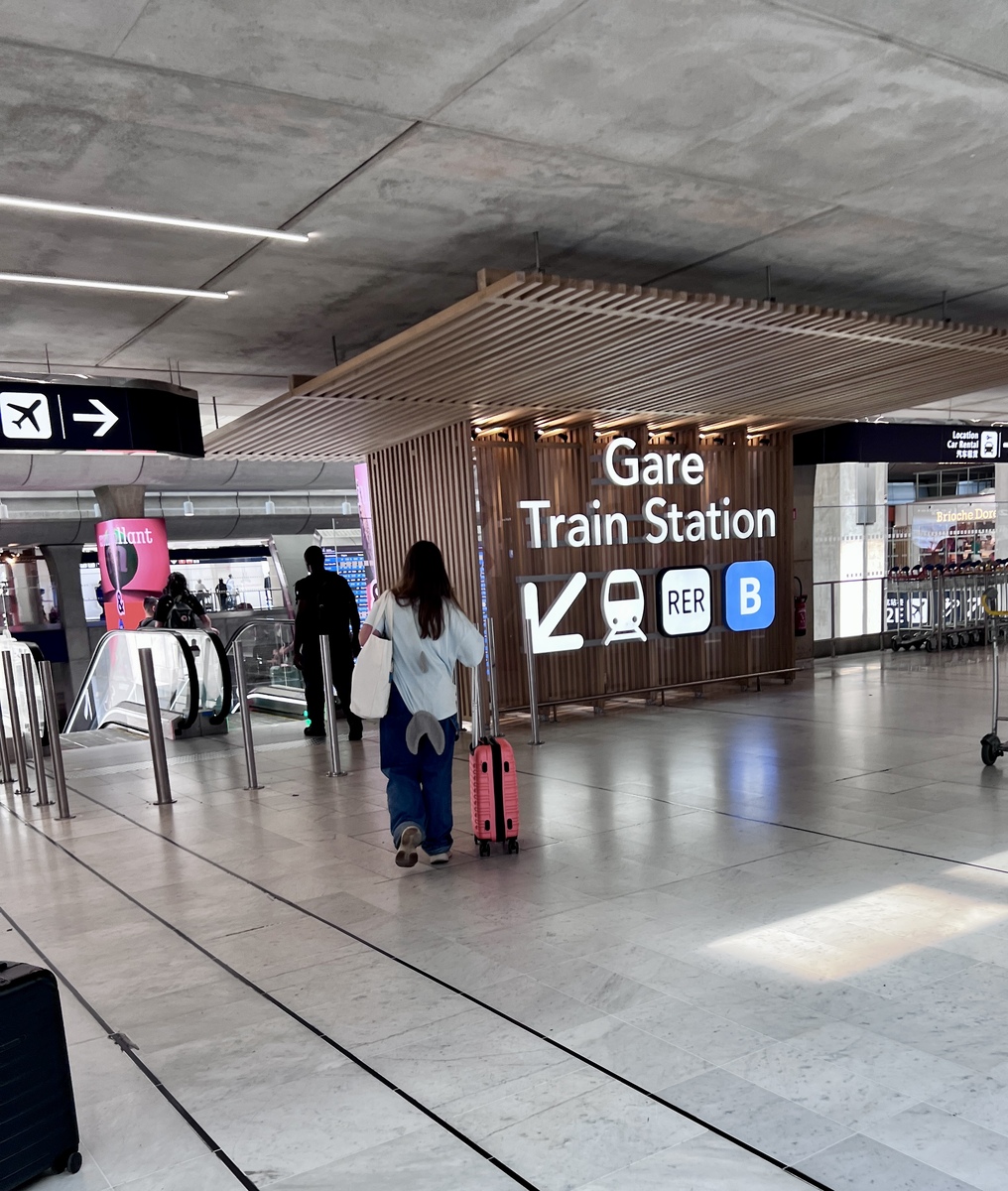 A traveler walks toward the RER B Gare train station sign in a modern, well-lit terminal. Escalators and directional signs are visible.