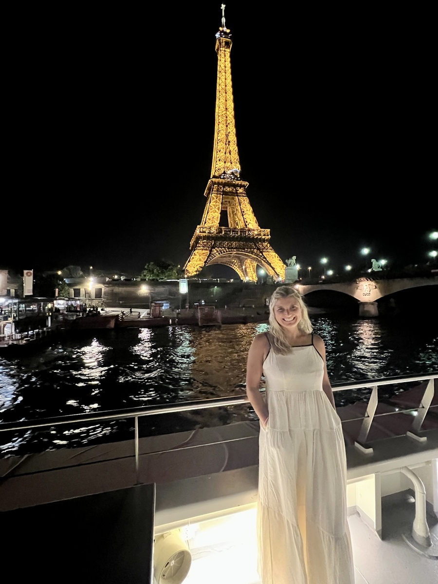 Brenda in a white dress smiles on a boat with the Eiffel Tower illuminated in the background against a night sky. The Seine River reflects lights, creating a serene, romantic atmosphere.