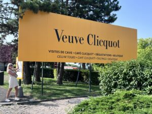 A woman in a white dress and sunglasses stands smiling beside a large orange "Veuve Clicquot" sign amidst green shrubs and trees, exuding a cheerful, summery vibe.