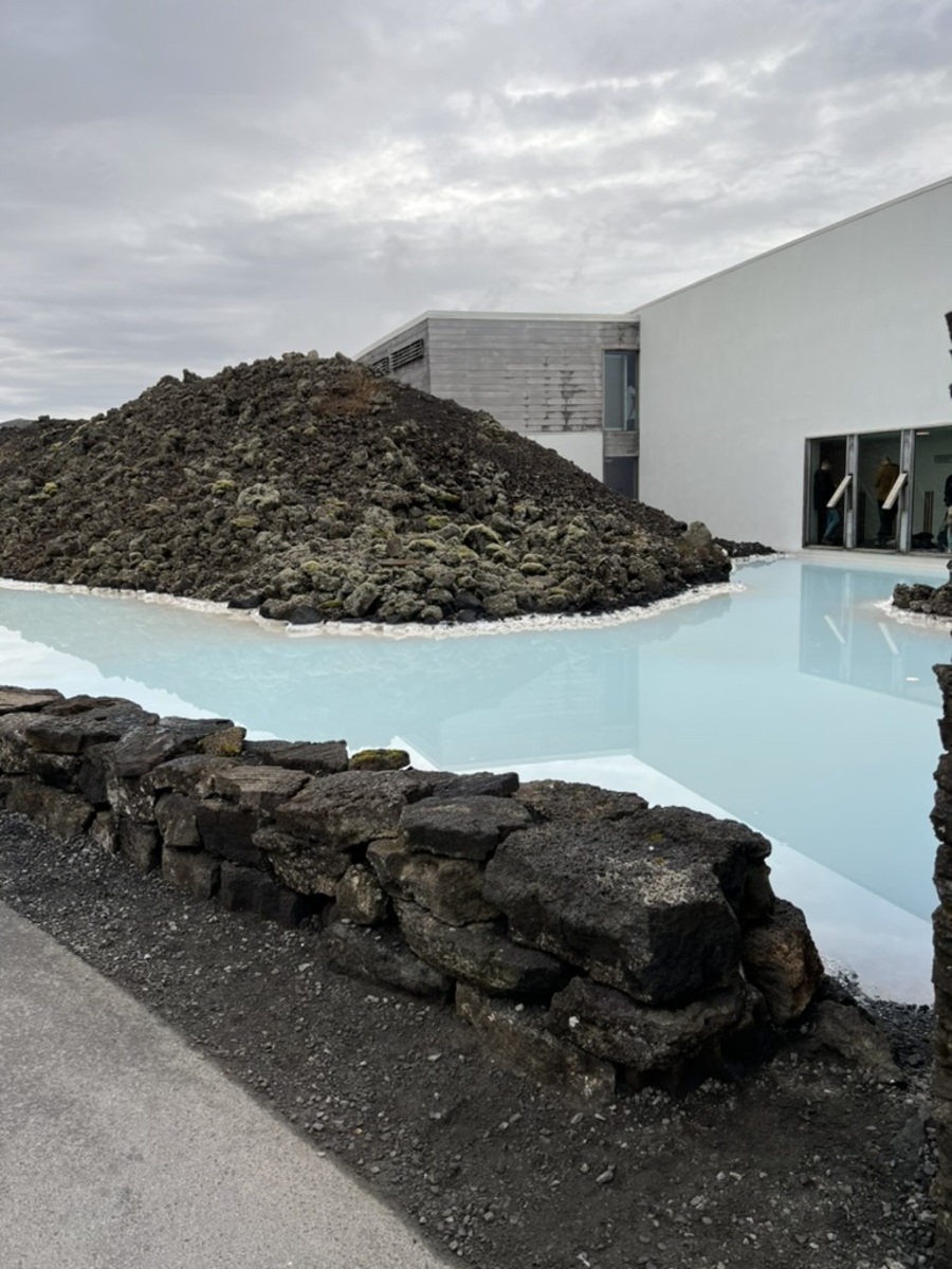 A tranquil, pale blue lagoon contrasts with rugged, dark volcanic rocks and modern white building under an overcast sky, creating a serene environment.