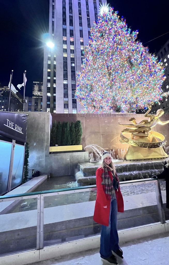 Brenda in a red coat and scarf stands smiling in front of a large, brightly lit Christmas tree at an ice rink. The scene conveys festive joy.