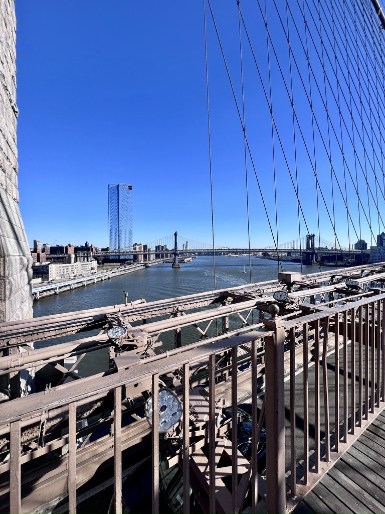 View from Brooklyn Bridge featuring its cables and beige railings, overlooking the East River with city skyline and blue sky, conveying a sense of openness.
