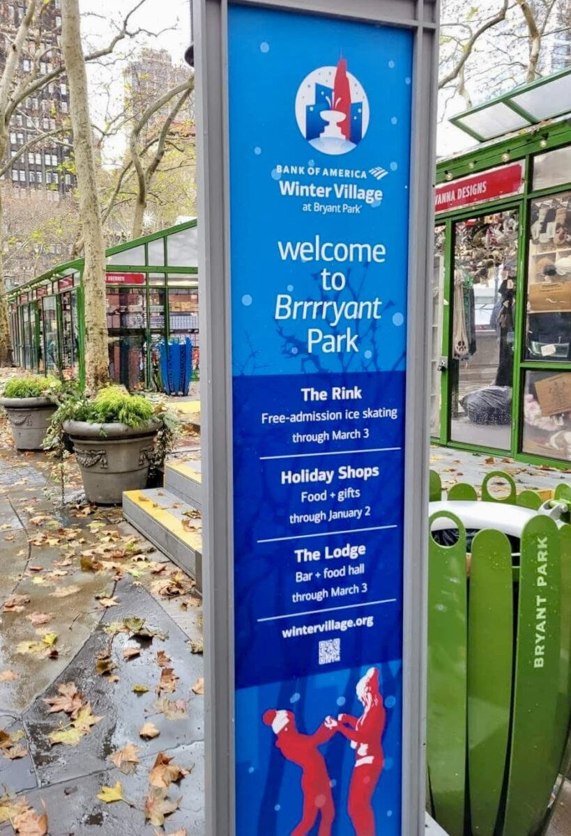 Tall blue sign at Bryant Park advertises "Winter Village" with ice skating, holiday shops, and food. Surrounding trees and leaves create a festive atmosphere.