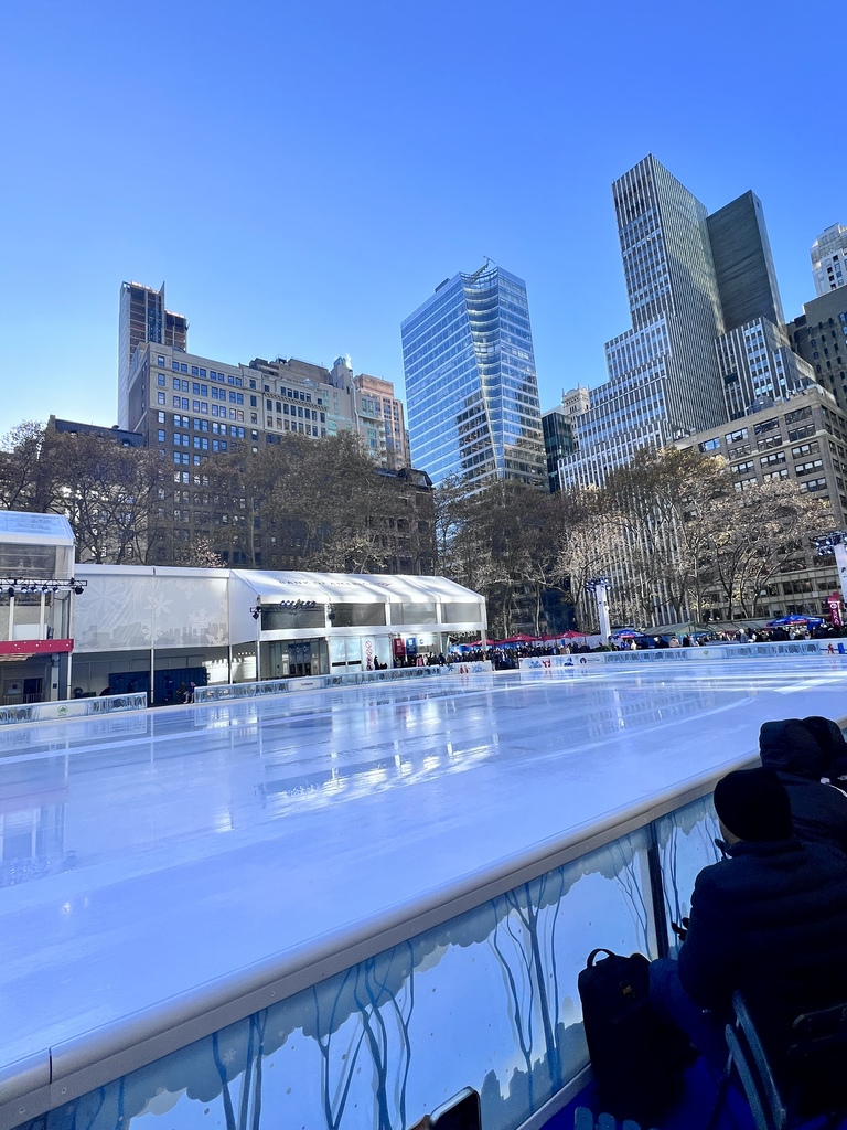 Ice rink with clear blue skies in an urban setting, surrounded by tall skyscrapers and leafless trees. People sit by the rink’s edge, creating a lively, winter atmosphere.