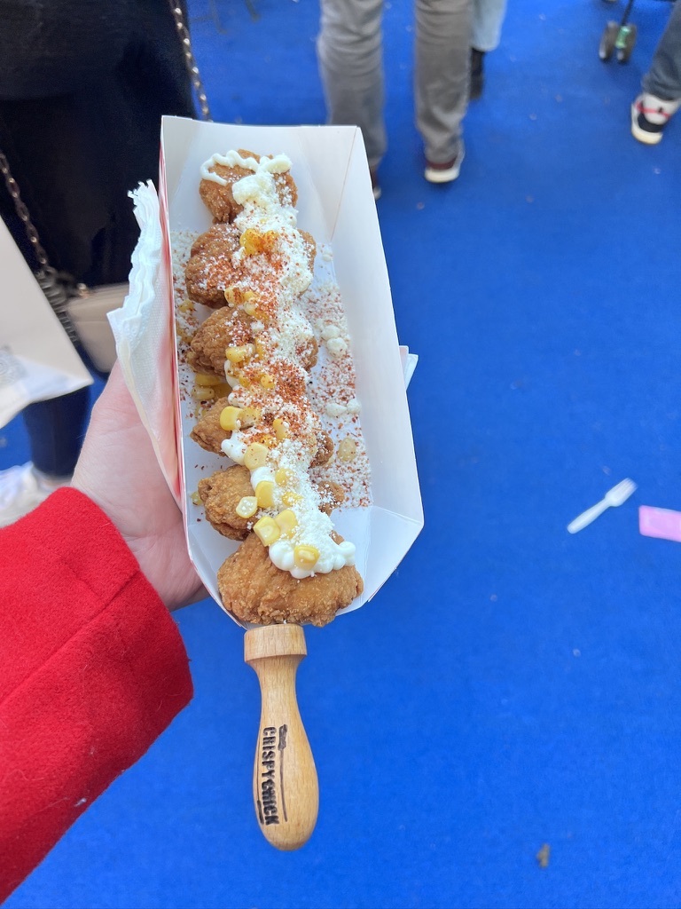 Hand holding a skewer of fried chicken nuggets topped with mayo, cheese, and corn in a paper tray. Set against a bright blue ground with people in the background.
