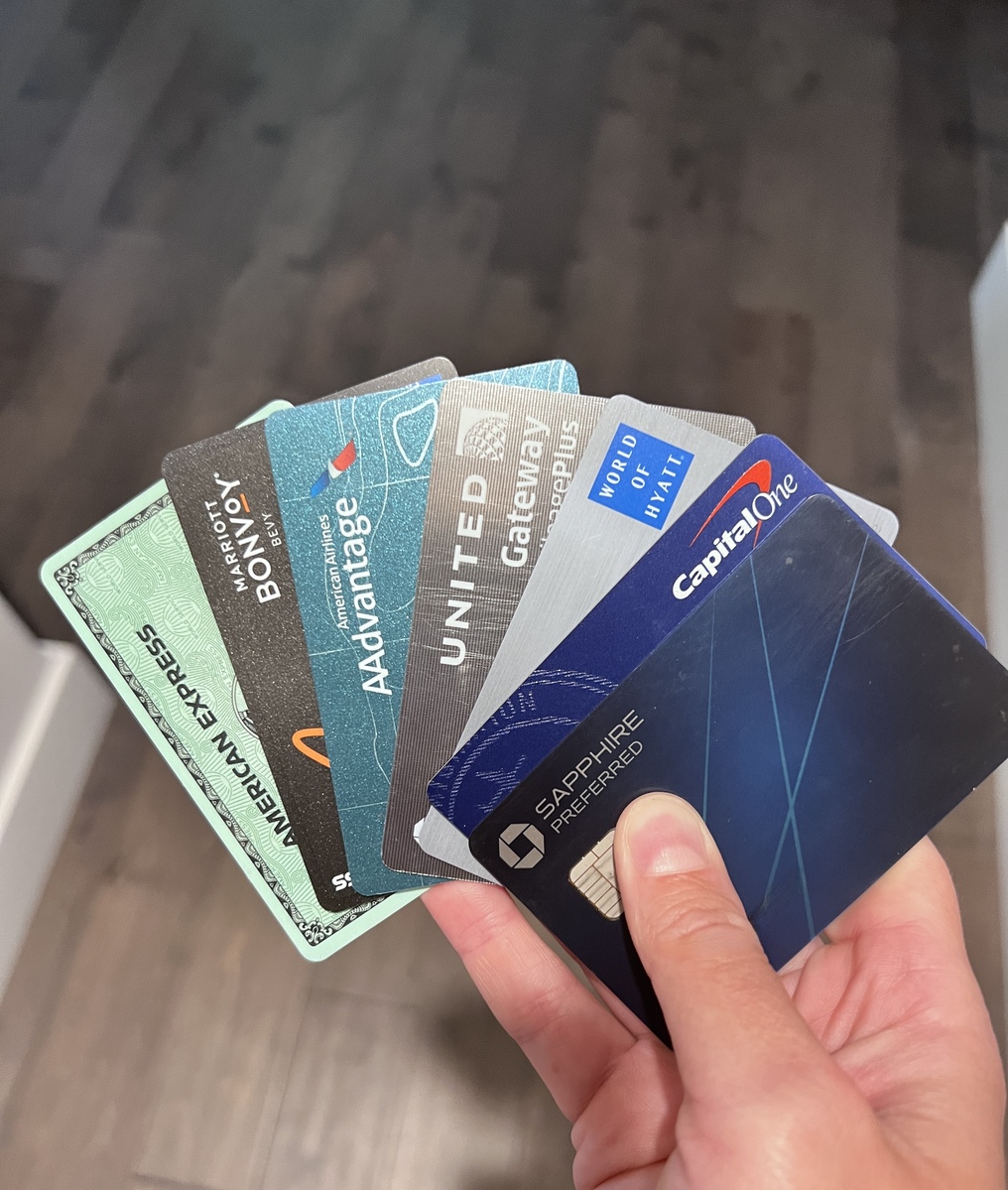 A hand fan holds six different credit cards, including American Express, Capital One, and Chase Sapphire Preferred, against a wooden floor backdrop.