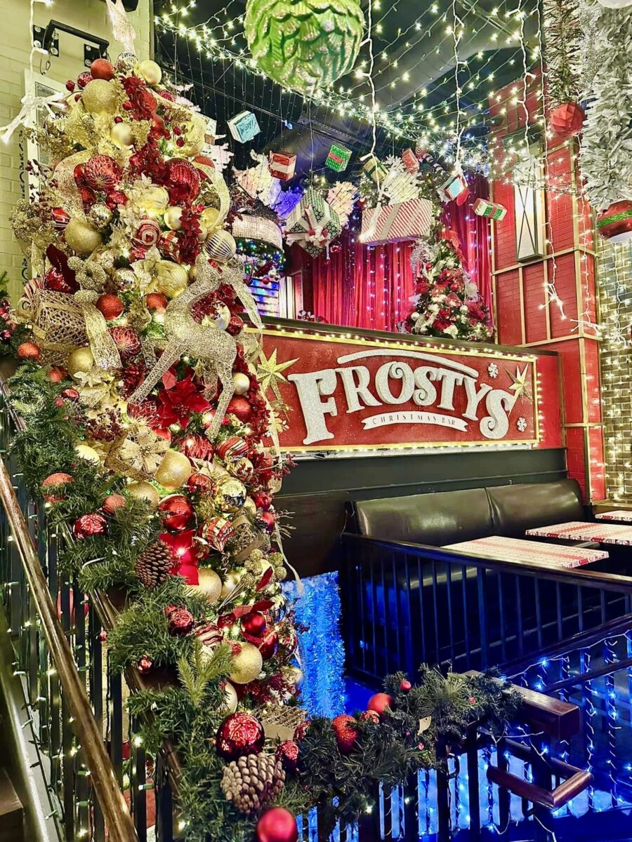 Festive bar scene with "Frosty's" sign, decorated with red and gold ornaments, reindeer, and lights. Cozy and cheerful Christmas atmosphere.