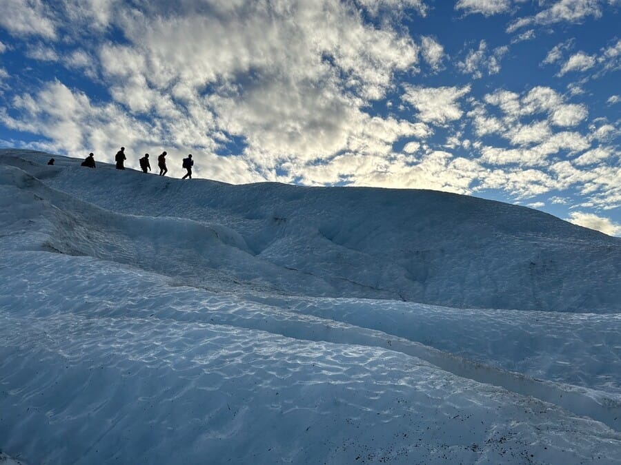 Silhouettes of hikers walk along a blue ice glacier under a sky filled with scattered clouds, evoking a sense of adventure and exploration.
