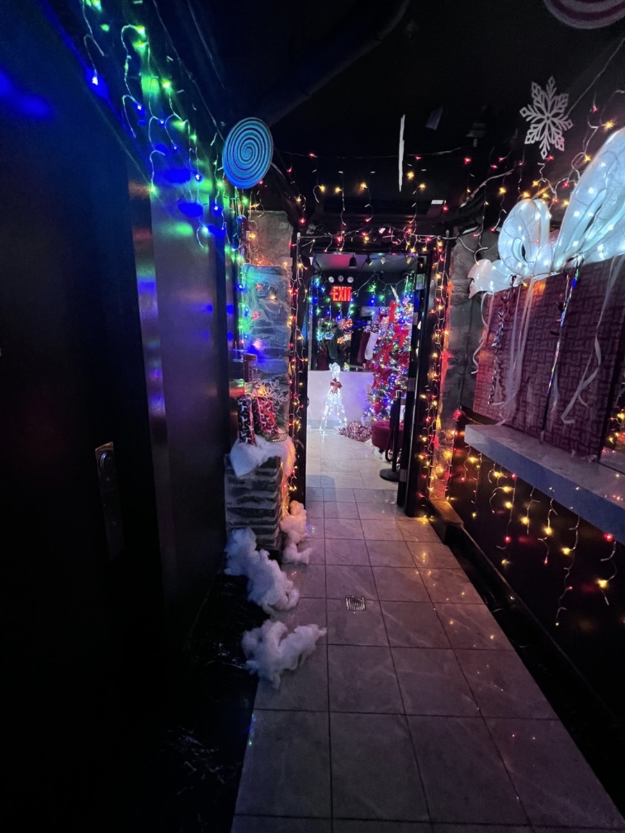 A hallway adorned with colorful Christmas lights leads to a festively decorated room. Snow-like decorations and hanging ornaments create a joyful, holiday ambiance.