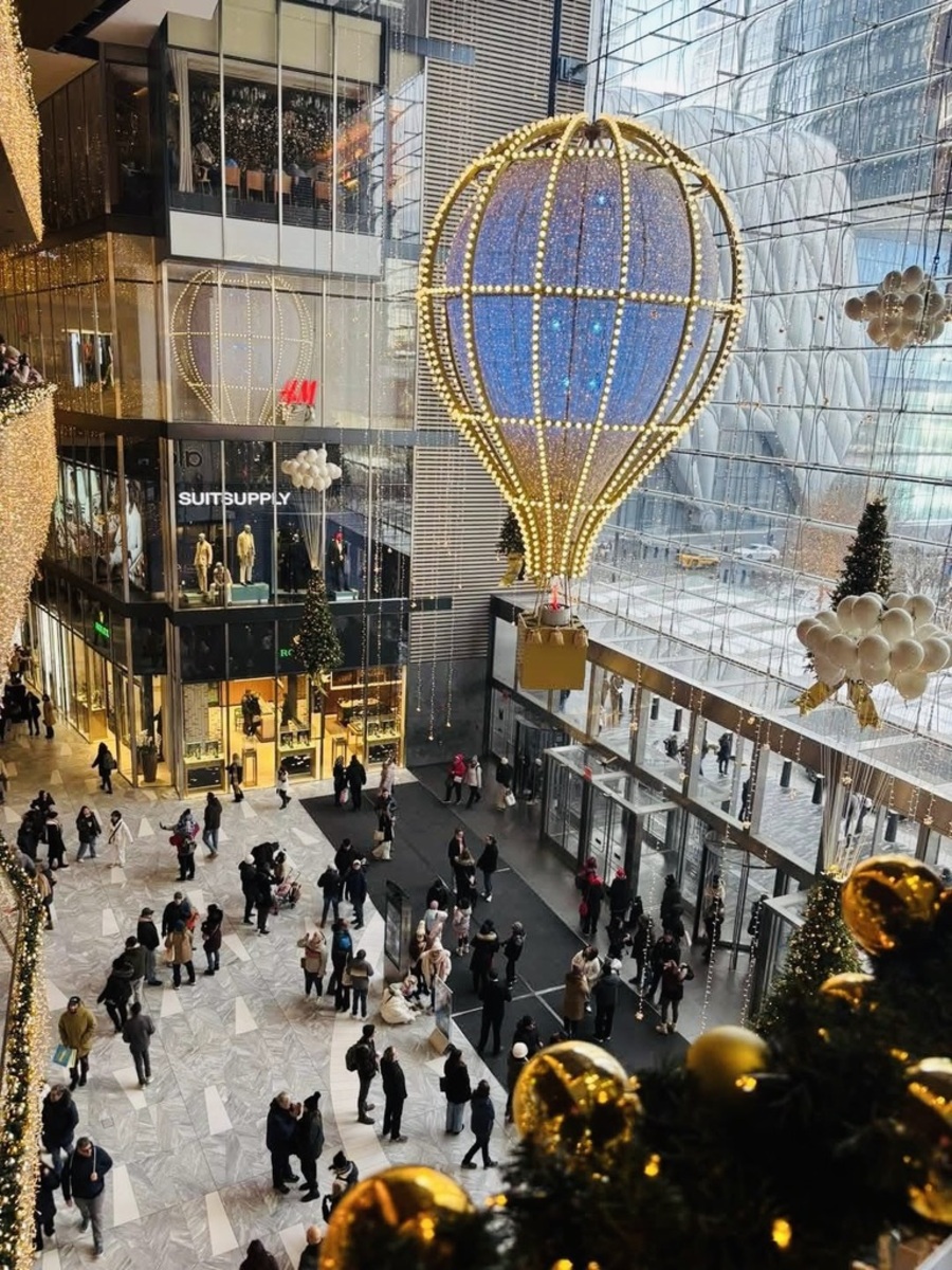Shopping mall atrium adorned with holiday decorations, including a large illuminated hot air balloon. Shoppers walk below on a busy, festive floor.