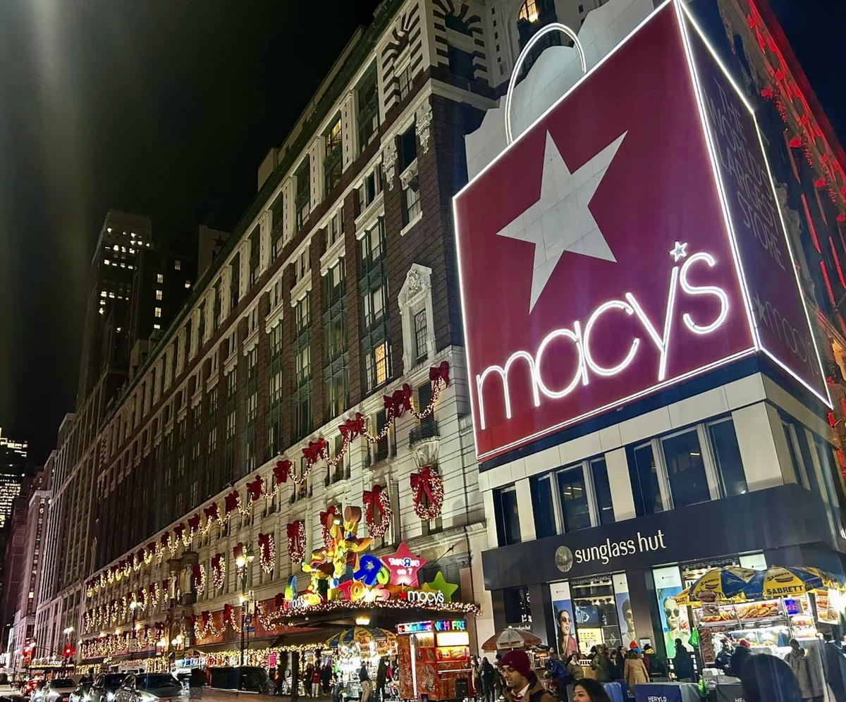 Festive night scene of a city street with a large Macy's sign and holiday decorations. The building is adorned with wreaths and lights, creating a joyful atmosphere.
